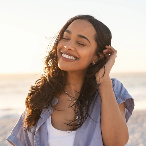 A smiling woman with long curly hair stands on the beach.